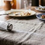 Saleiro de vidro tombado sobre toalha de linho, com uma linha de sal derramado na mesa do almoço