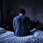 Pessoa sentada na beira da cama de madrugada, de costas, com a camiseta úmida de suor